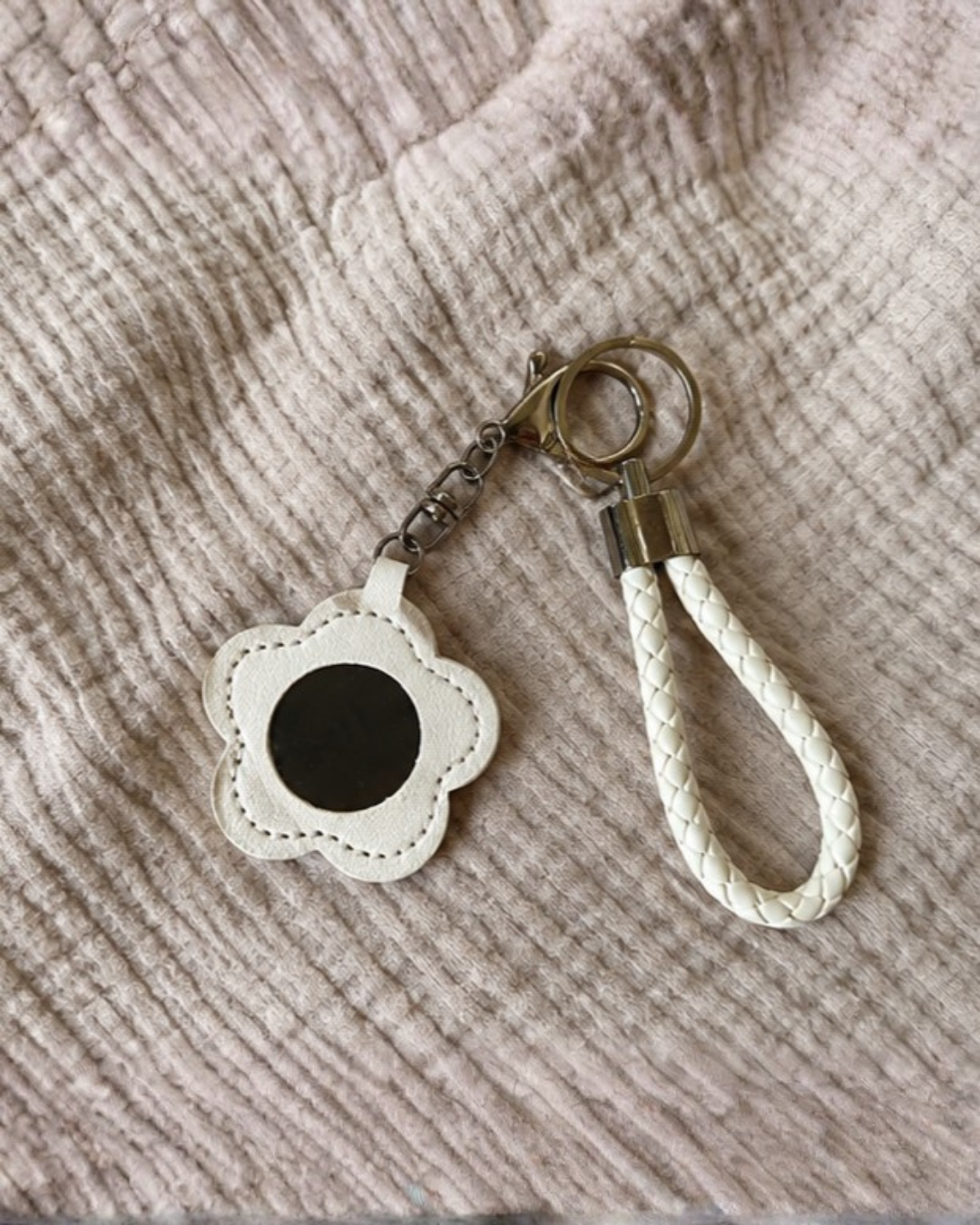 A white faux leather flower-shaped keychain with a black center and a white braided loop, resting on a textured beige fabric background.