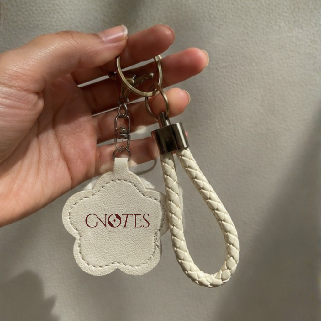 A hand holding a cream-colored faux leather keychain with a cloud-shaped tag printed with 'GNOTES' in burgundy and a matching braided cream key strap with a silver metal clasp.