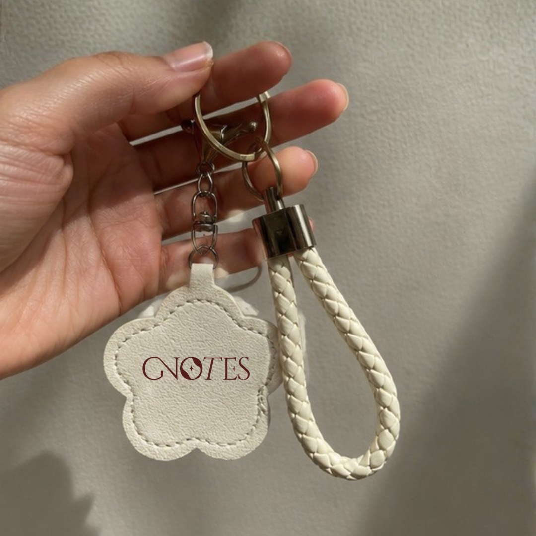 A hand holding a cream-colored faux leather keychain with a cloud-shaped tag printed with 'GNOTES' in burgundy and a matching braided cream key strap with a silver metal clasp.
