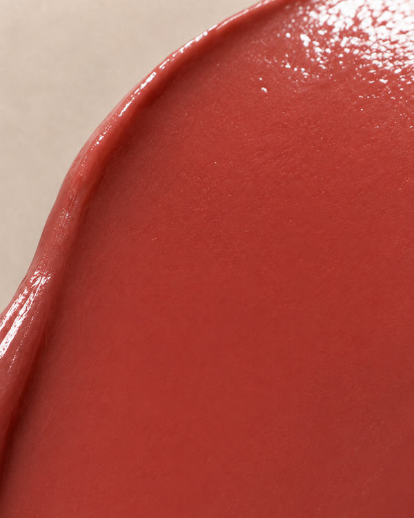 A close-up texture shot of a glossy, smooth cosmetic cream or lip gloss in a warm coral-red shade, with a curved edge showing the product's creamy consistency against a neutral beige background.