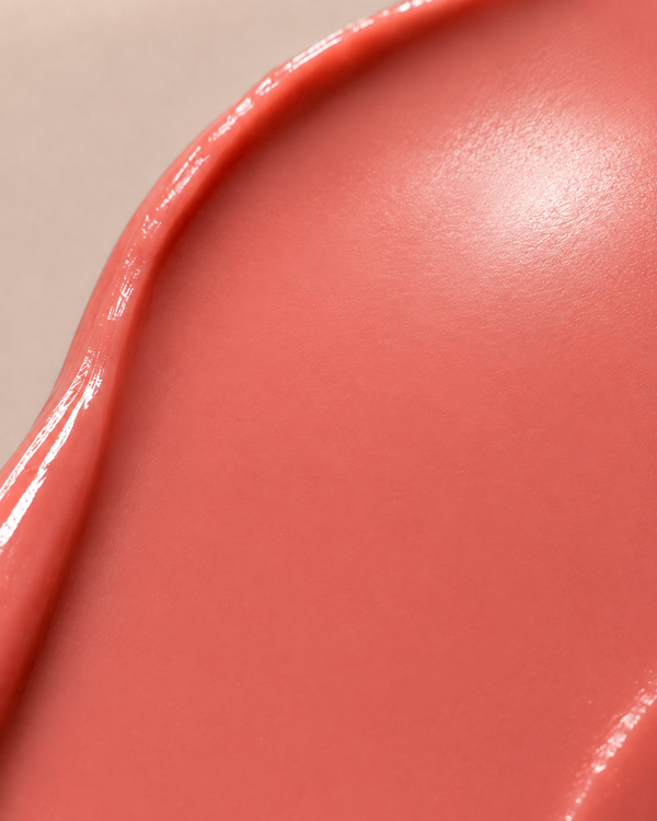 A close-up of a glossy, smooth swatch of coral pink cosmetic cream or gel, showing a subtle sheen and texture against a light neutral background.